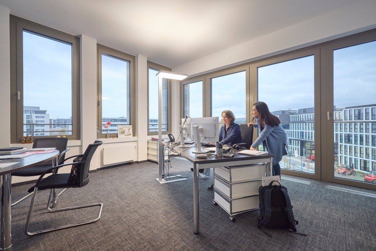 Zwei IQWiG Mitarbeiterinnen an einem Schreibtisch in einem Büro mit Fensterfront